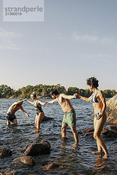 Multiracial male and female friends holding hands while standing in lake against sky