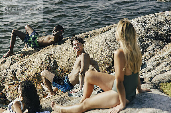 Smiling shirtless man talking with female friend sitting on rock near lake