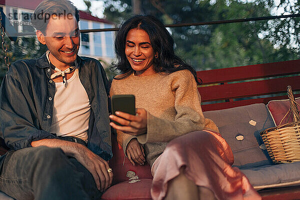 Smiling woman sharing smart phone with male friend while sitting on bench in back yard