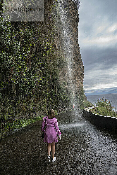 Woman stands watching a waterfall splashing down on a coastal roadway  an extreme natural waterfall car wash in the rocks of Madeira  Portugal; Canhas  Madeira  Portugal