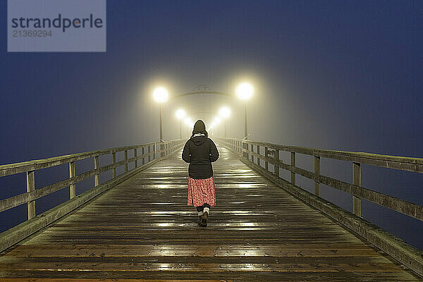 View taken from behind of a woman walking on a wooden pier at night at White Rock; British Columbia  Canada