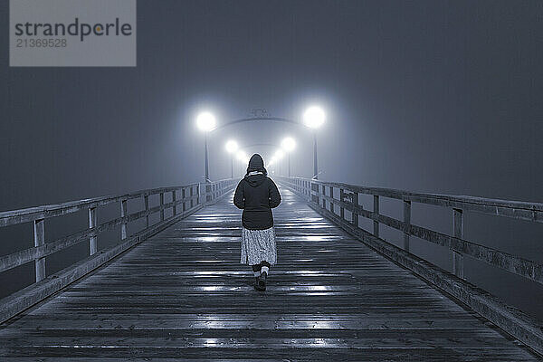 View taken from behind of a woman walking on a wooden pier at night at White Rock; British Columbia  Canada