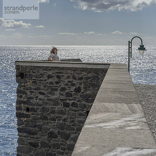 Woman stands along a stone wall along the waterfront of Ponta do Sol on the coast of Portugal  with the ocean water shimmering in the sunlight; Ponta do Sol  Madeira  Portugal