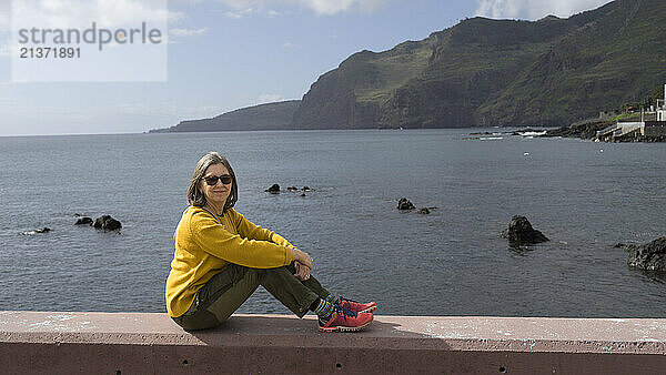 Woman poses for the camera as she sits on a concrete wall relaxing in the warm sun at the water's edge along the coast of the island of Madeira  Portugal; Canical  Madeira  Portugal