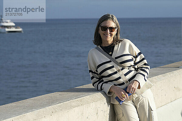 Senior woman with sunglasses poses along a concrete wall in the sunlight along a waterfront on the island of Madeira  Portugal; Funchal  Madeira  Portugal