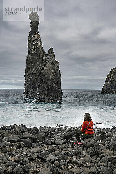 Woman sits on the rocks along the shore viewing the Ilheus da Ribeira da Janela rock formation along the coast of the island of Madeira in Portugal; Ribeira da Janela  Madeira  Portugal