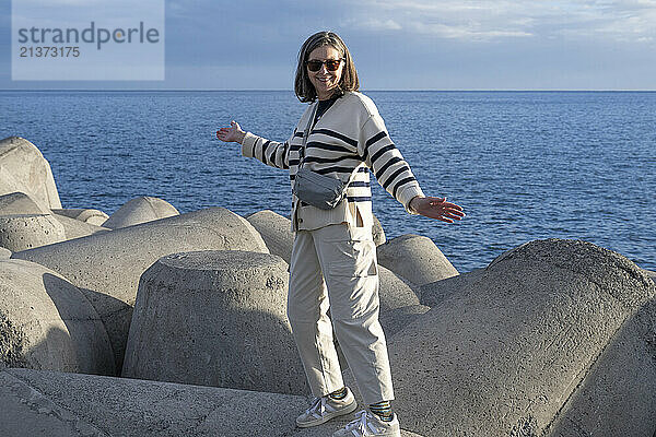 Senior woman with sunglasses poses in the sunlight along concrete structures on the waterfront of Funchal on the island of Madeira  Portugal; Funchal  Madeira  Portugal