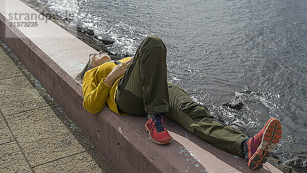 Woman lays on her back on a low concrete wall relaxing in the warm sun at the water's edge along the coast of the island of Madeira  Portugal; Canical  Madeira  Portugal