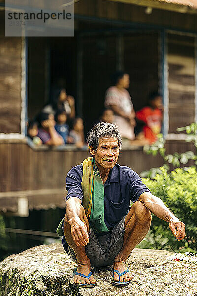 Mature man crouches on a boulder  with people on a balcony in the background; Rante Pao  North Toraja  South Sulawesi  Indonesia