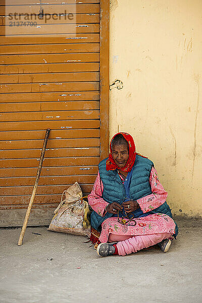 Woman sits on the ground against a building wall  with a bag and walking stick beside her; Spiti Valley  Himachal Pradesh  India