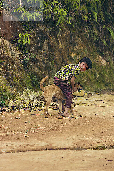 Boy sits and plays on the back of his pet dog; Ban Nam Nghiep  Muong La  Son La  Vietnam