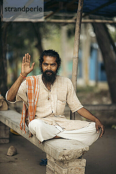 Portrait of a mid adult man in traditional indian clothing sitting cross-legged on a concrete bench at the Lower Ganga Canal bridge in India; India