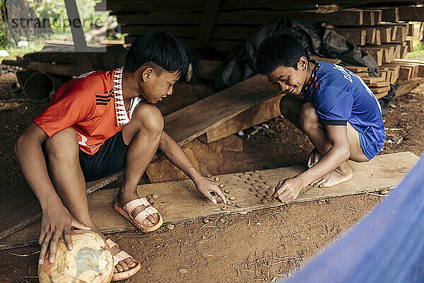 Two boys enjoy playing a game with shells on the ground in a village in Laos  both wearing athletic clothing and holding a soccer ball; Kok Phung Tai  Champasak Province  Laos