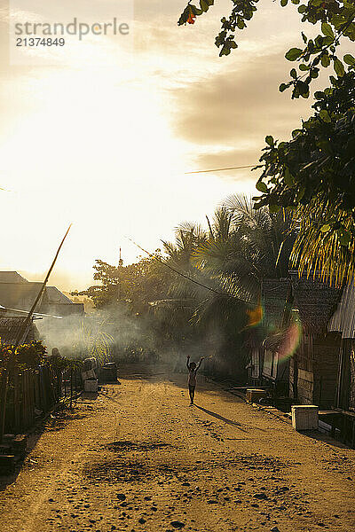 Woman stands with her arms raised into the water's mist which creates a rainbow in a tropical community setting; Lambako  Banggai Laut  Central Sulawesi  Indonesia