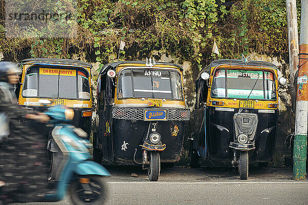 Auto rickshaws parked in a row along a street with a woman riding by on a motor scooter  in Jammu  India; Jammu  Jammu And Kashmir  India