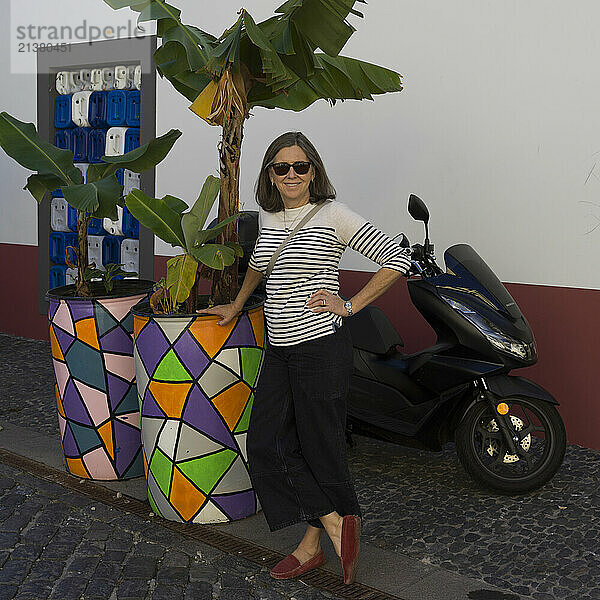 Woman with sunglasses stands posing for the camera  leaning on a colourful planter on a street with a motor scooter parked behind her  in Camara de Lobos on the island of Madeira  Portugal; Camara de Lobos  Madeira  Portugal