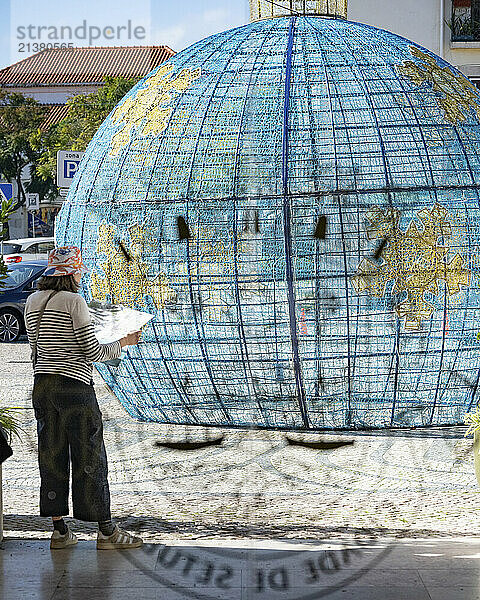 Woman stands reading a brochure in front of a sculpture of a large decorative Christmas bauble sculpture; Setubal  Lisbon  Portugal