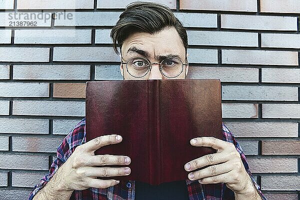 Funny face. Young hipster guy wearing eyeglasses with picked up one browe looking at camera and holding a book or note book on brick wall background