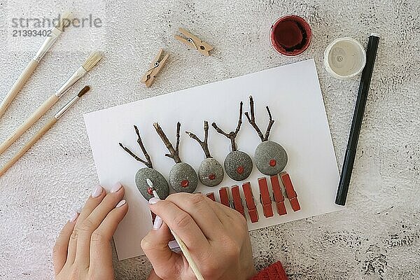 Step by step. Top view. Process kid children craft. Do it yourself Diy Merry Christmas deer on greeting card postcard made of pebbles  sea stones  clothespins and branches on white background. Natural Eco style diy. Gift idea