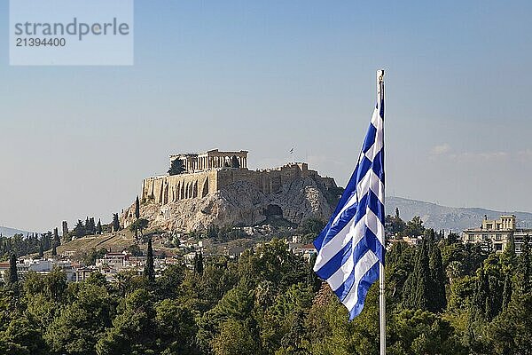 A picture of the Acropolis of Athens  and the Parthenon  as seen above a tree line and the Greek flag in the foreground