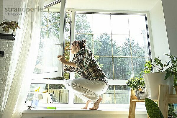 Woman manually washes the window of the house with a rag with spray cleaner and mop inside the interior with white curtains. Restoring order and cleanliness in the spring  cleaning servise