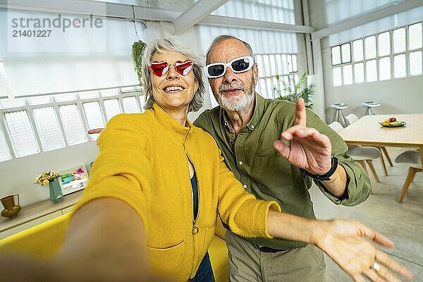 Senior woman with heart shape sunglasses taking selfie with husband while having fun at home