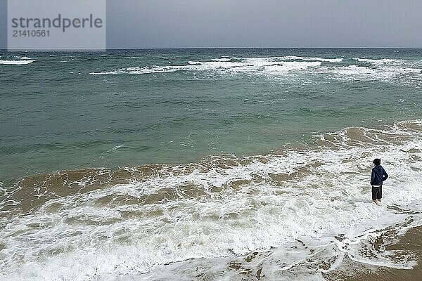 Drone aerial of young boy in shorts standing on the summer beach and looking at the stormy spindrift waves. View from the back. Stormy weather on the sea coast. Stormy summer day