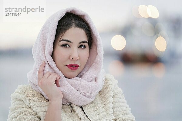 Winter portrait of brunette with red plump lips  dressed in white fur coat and light pink scarf on head. Soft lighting on elegant young adult female model on evening. Depth of field  selective focus