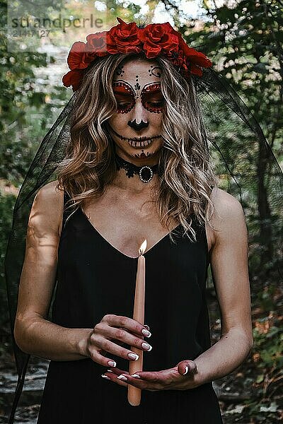 Happy Halloween concept. Scared horrible female in special outfit  wears white clay skull  professional sugar skull makeup to look spooky  wears black clothes playing with candle. Day of The Dead. Dia de los muertos