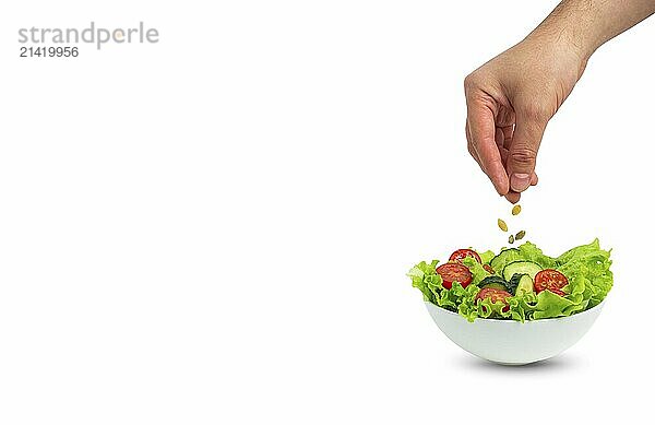 White bowl of salad with fresh cucumbers  tomatoes  iceberg salad leaves and male hand sprinkling pumpkin seeds over salad isolated on green background  template for advertising  space for text  food photography