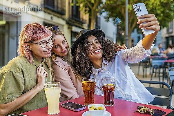 Happy female friends taking selfie together sitting on a sidewalk cafeteria drinking soda and eating snacks