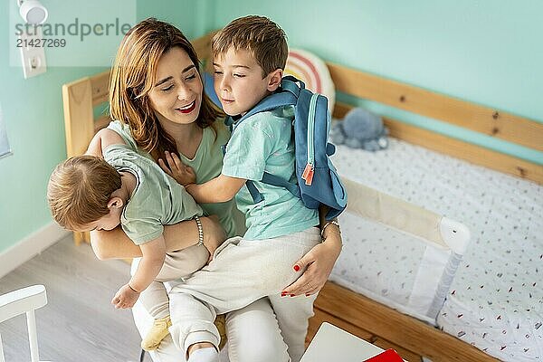 Mother embracing and holding a school boy and baby sitting together in the bedroom before going to school