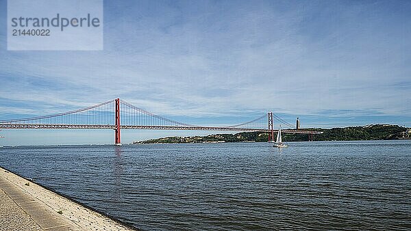 View of a red bridge over a calm river with a sailing boat and promenade  Ponte 25 de Abril  Lisbon  Portugal  Europe