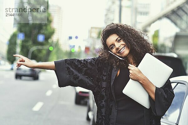 Young happy black girl try to take a taxi on the street holding a laptop