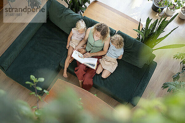 Mature woman reading book to daughters on sofa in living room at home