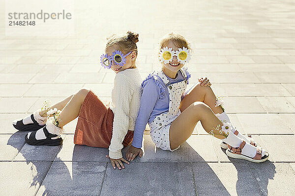 Smiling friends with flower sunglasses sitting on footpath