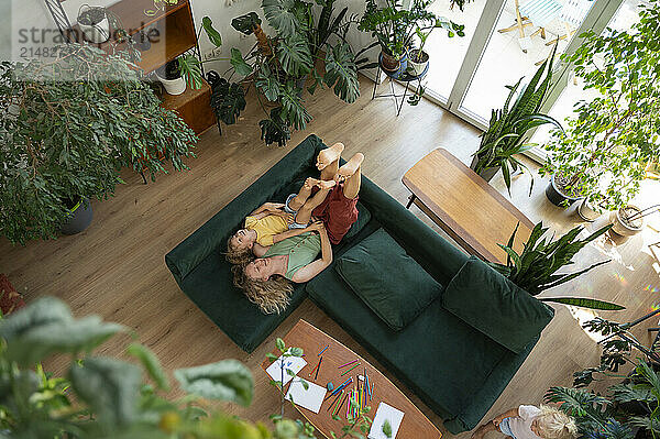 Happy mother and daughter lying on sofa amidst plants in living room at home