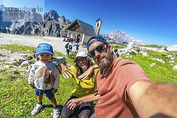 Family taking a selfie with the stunning tre cime di lavaredo mountains and chapel in the background  enjoying summer vacation in the dolomites. Cappella degli Alpini