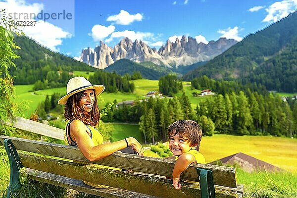 Mother and son relaxing on a bench enjoying the landscape of the odle mountains in val di funes in the dolomites