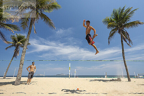 Trickliner jumping over slackline between coconut palm trees in Ipanema Beach boardwalk shore  on a sunny day with blue sky  Rio de Janeiro  Brazil