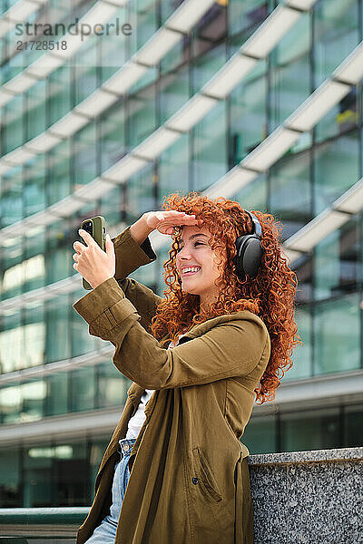 Stylish redhead woman with headphones using mobile phone and shielding her eyes from the sun in front of modern building