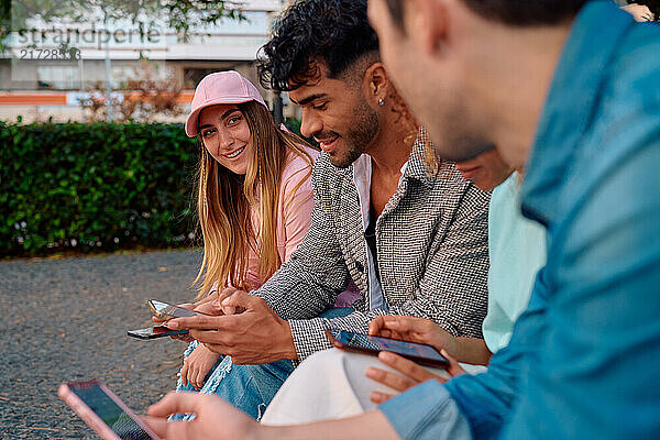 Four friends are sitting on a bench in a park  each focused on their smartphones. They are outdoors  surrounded by greenery  enjoying their time together in a relaxed atmosphere.