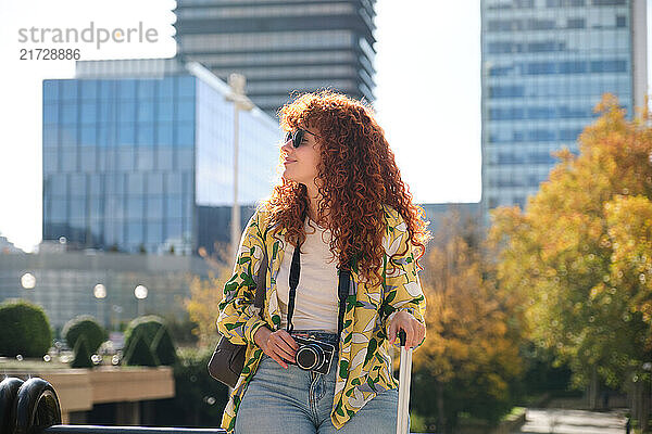 Young woman with red hair  camera and suitcase exploring a modern city during vacation