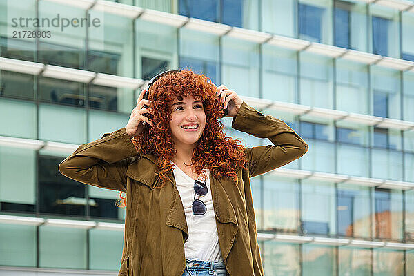 Happy redhead woman enjoying music with headphones in front of modern building