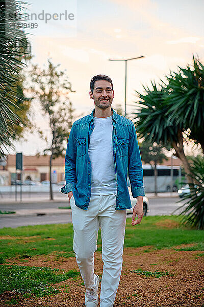 A young man strolls through a park surrounded by greenery and palm trees at sunset. He wears a denim shirt over a white tee and white pants  exuding a casual  relaxed vibe.