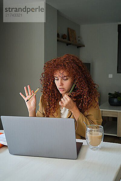 Young woman with curly red hair gesturing with pens while using laptop and talking on the smartphone at home office