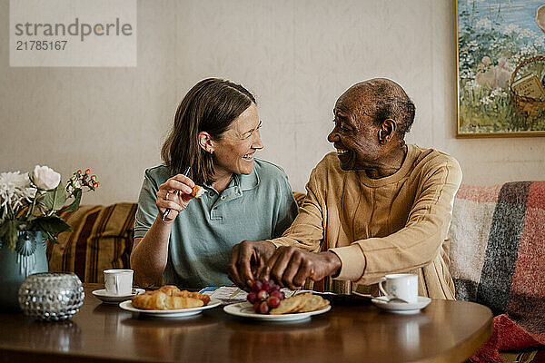 Happy female nurse talking with elderly male while having breakfast at home