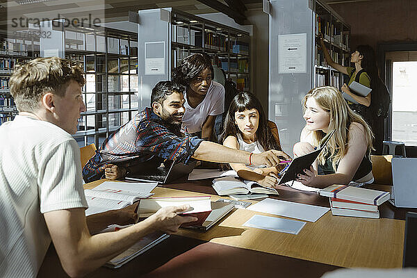 Smiling man pointing at book while doing group study with male and female friends in library at university