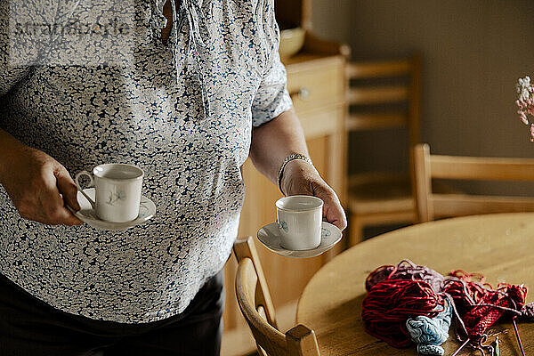 Senior woman holding saucers with tea cups near dining table at home