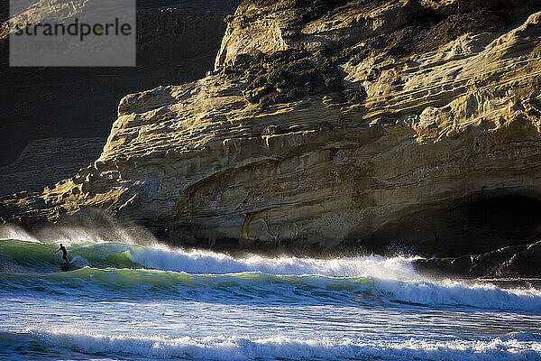 Unidentified surfer riding a wave under a enormous cliff. Cold  Oregon surf conditions attest to the devotion of the surfer.